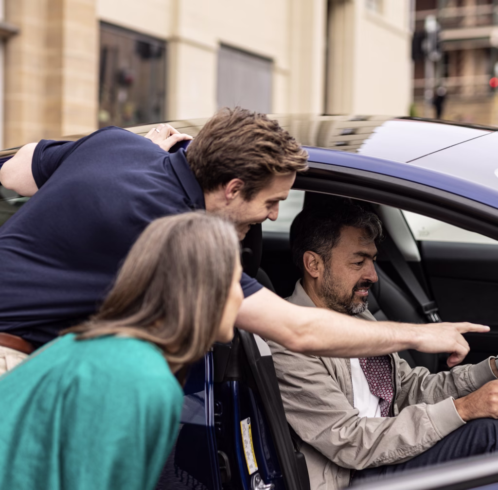 A Drive Car Hire employee shows two people how to find something in their Tesla's settings. He points at a screen just out of view as a man sits in a blue Tesla and a woman looks on.